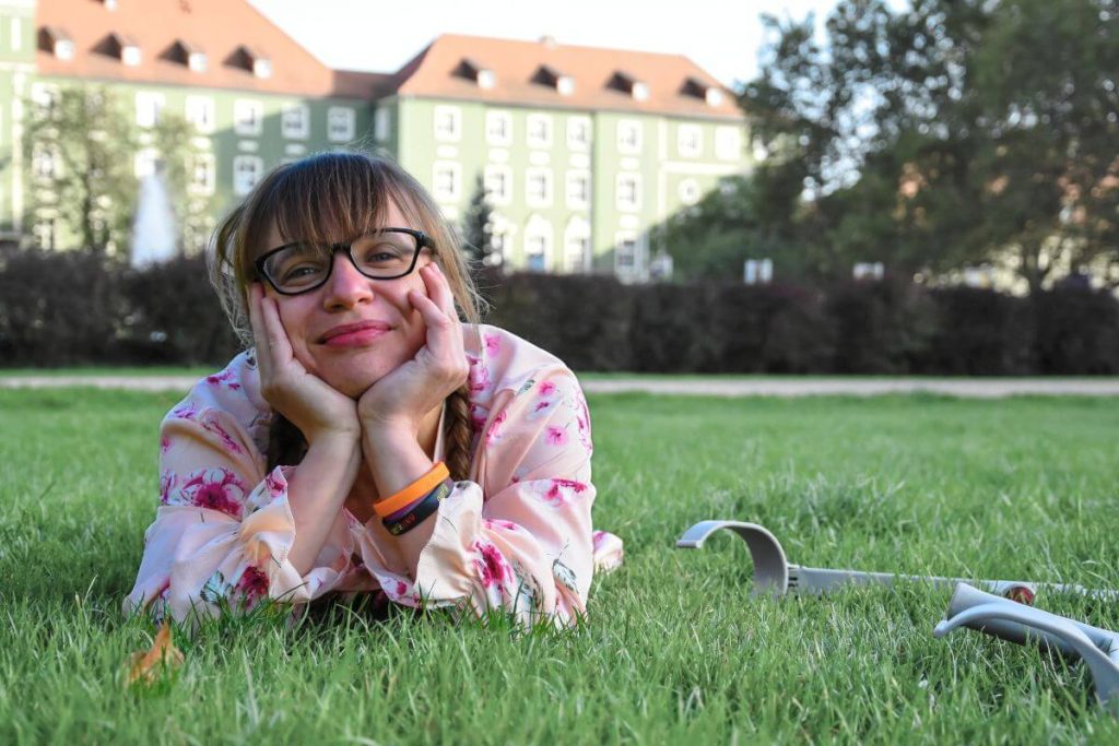 Jagoda Gmachowska - młoda uśmiechnięta kobieta w okularach leży na trawie podpierając głowę obiema rękami, patrzy w stronę obiektywu, włosy zaplecione w dwa warkocze z grzywką, widoczne są rękawy kremowej sukienki w różowe kwiaty, obok na trawie leżą kule łokciowe; w tle, za żywopłotem widoczny duży zielony budynek z czerwonym dachem