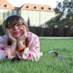 Jagoda Gmachowska - młoda uśmiechnięta kobieta w okularach leży na trawie podpierając głowę obiema rękami, patrzy w stronę obiektywu, widoczne sąrękawy kremowej sukienki w różowe kwiaty, obok na trawie leżą kule łokciowe; w tle, za żywopłotem widoczny duży zielony budynek z czerwonym dachem